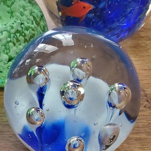 May include: A selection of glass paperweights. One has a blue and white design with silver bubbles. Another features a red fish in a blue and yellow underwater scene. A third paperweight is green.
