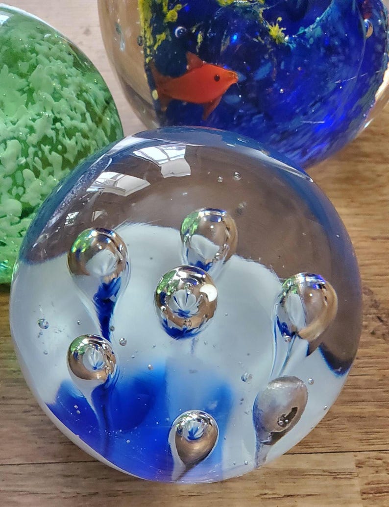 May include: A selection of glass paperweights. One has a blue and white design with silver bubbles. Another features a red fish in a blue and yellow underwater scene. A third paperweight is green.