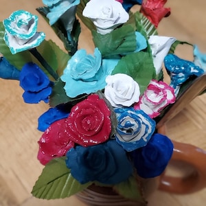 May include: A bouquet of paper flowers in a brown ceramic vase. The flowers are painted in shades of blue, red, and white.
