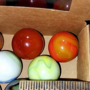 May include: A collection of colorful glass marbles in a cardboard box. The marbles vary in color, including red, orange, green, and white, with swirled and solid patterns. A wooden ruler is visible in the foreground.