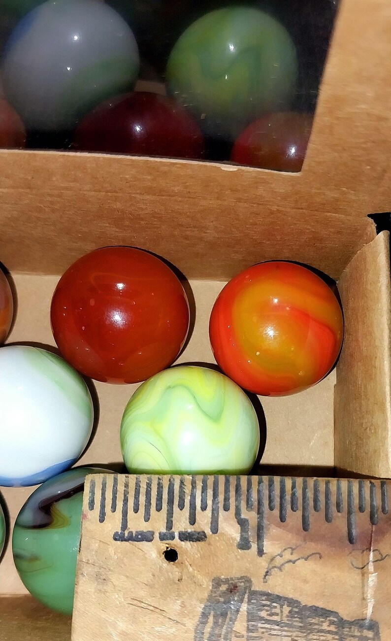 May include: A collection of colorful glass marbles in a cardboard box. The marbles vary in color, including red, orange, green, and white, with swirled and solid patterns. A wooden ruler is visible in the foreground.