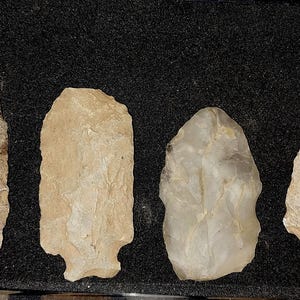 May include: Four arrowheads made of stone, displayed on a black background. The arrowheads are various shades of white and gray.
