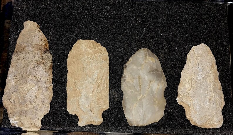 May include: Four arrowheads made of stone, displayed on a black background. The arrowheads are various shades of white and gray.