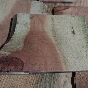 May include: Close-up of several pieces of unfinished wood. The wood has a natural grain pattern with shades of red, brown, and tan. Some pieces have a curved edge. The wood appears to be ready for crafting or woodworking projects.
