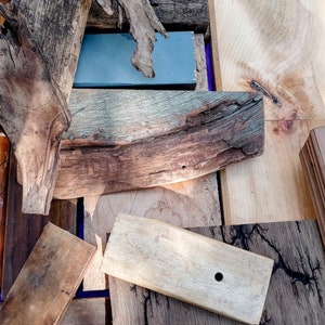 May include: A collection of wood pieces, including a piece of driftwood, a piece of wood with a hole drilled in it, and a piece of wood with a dark brown stain. The wood pieces are arranged in a random pattern on a wooden surface.