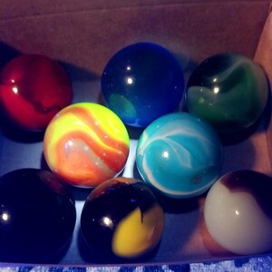 May include: A collection of colorful glass marbles in a cardboard box. The marbles are various colors, including red, blue, green, yellow, and white, with swirled and solid patterns. The box is lit with a blue light.