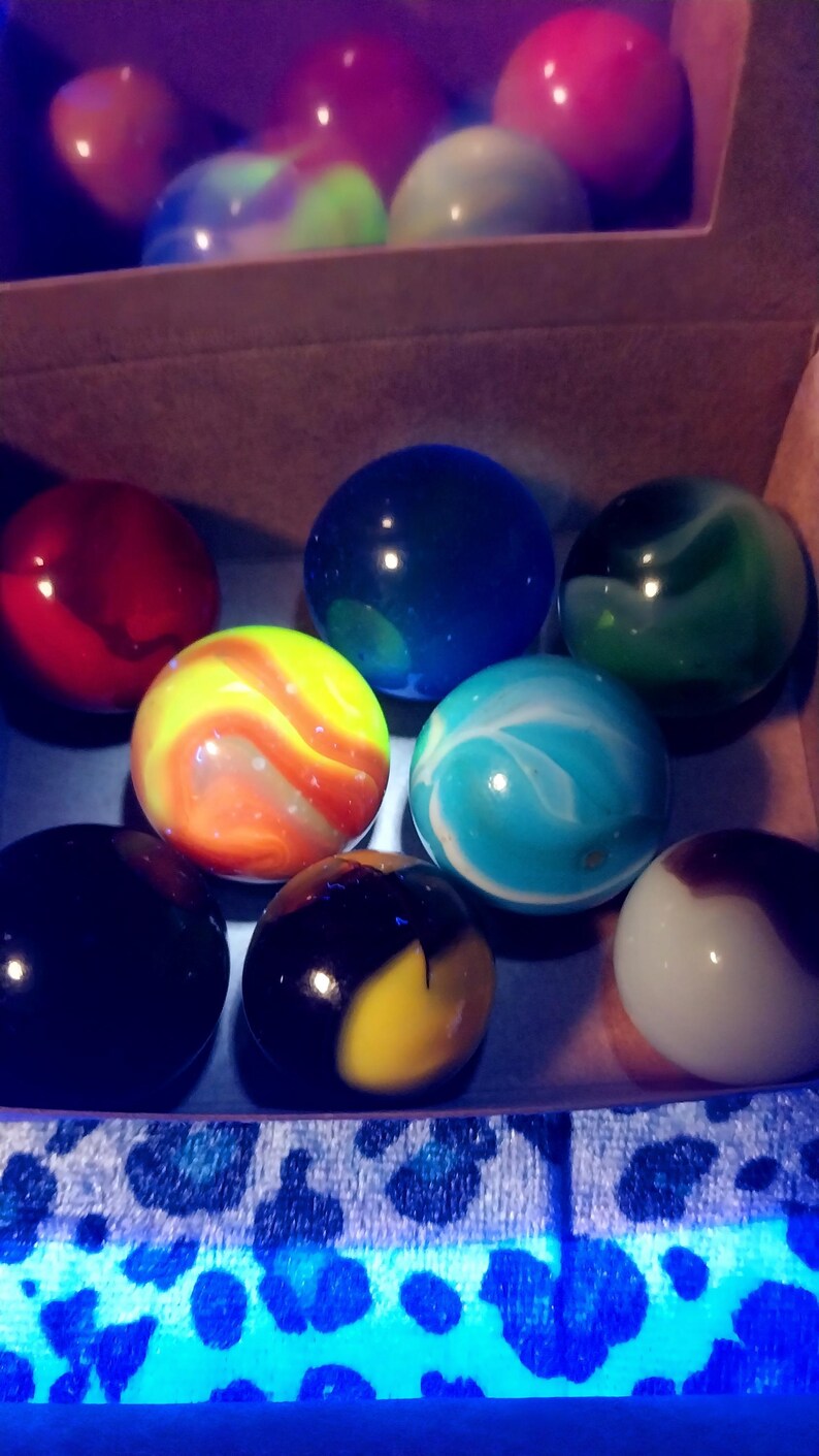 May include: A collection of colorful glass marbles in a cardboard box. The marbles are various colors, including red, blue, green, yellow, and white, with swirled and solid patterns. The box is lit with a blue light.