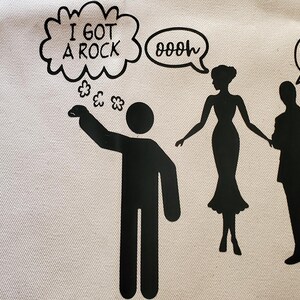 May include: A white tote bag with a black graphic of a person saying "I GOT A ROCK" with a thought bubble. Silhouettes of a woman and a man are also present. Various rocks are in the upper left corner.