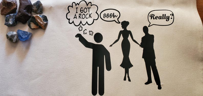 May include: A white tote bag with a black graphic of a person saying "I GOT A ROCK" with a thought bubble. Silhouettes of a woman and a man are also present. Various rocks are in the upper left corner.