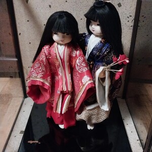 May include: Two Japanese dolls in traditional attire. The doll on the left wears a red kimono with gold accents, while the other doll is in a blue and gold kimono. Both dolls have black hair and are displayed on a black base.