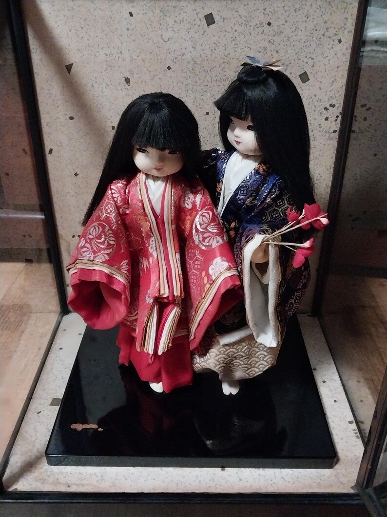 May include: Two Japanese dolls in traditional attire. The doll on the left wears a red kimono with gold accents, while the other doll is in a blue and gold kimono. Both dolls have black hair and are displayed on a black base.
