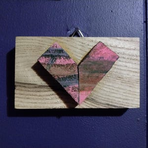 May include: A heart-shaped piece of art made from wood and painted in shades of pink, black, and brown. The heart is mounted on a wooden plaque.