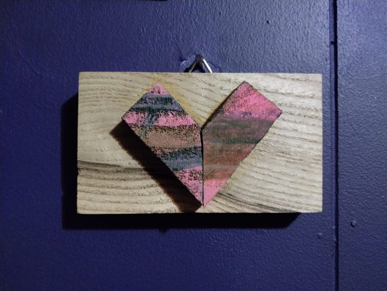 May include: A heart-shaped piece of art made from wood and painted in shades of pink, black, and brown. The heart is mounted on a wooden plaque.