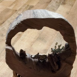 May include: A handcrafted wooden sculpture featuring a circular frame with a carved landscape. The sculpture includes miniature elephant figurines and plant details. The base is a woven basket.