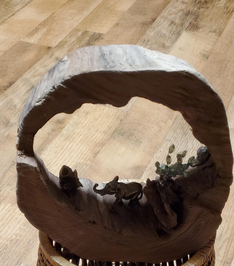 May include: A handcrafted wooden sculpture featuring a circular frame with a carved landscape. The sculpture includes miniature elephant figurines and plant details. The base is a woven basket.