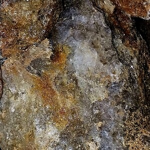 May include: Close-up of a rough, textured rock formation with a mix of colors. The rock features shades of brown, gold, gray, and white, with a crystalline structure visible in some areas. The surface has a rugged, natural appearance.