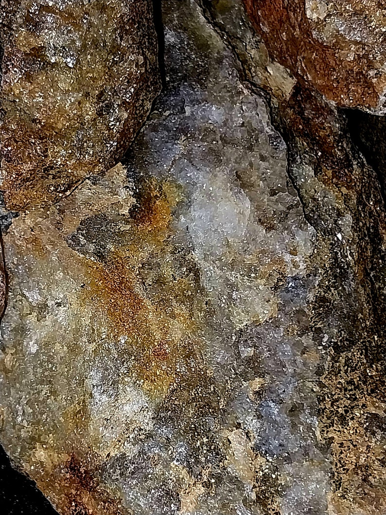 May include: Close-up of a rough, textured rock formation with a mix of colors. The rock features shades of brown, gold, gray, and white, with a crystalline structure visible in some areas. The surface has a rugged, natural appearance.