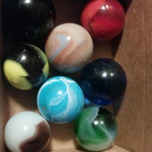 May include: A collection of colorful glass marbles in a cardboard box. The marbles are various colors, including black, red, blue, green, white, and a marbled blue and white. Some marbles have swirling patterns.