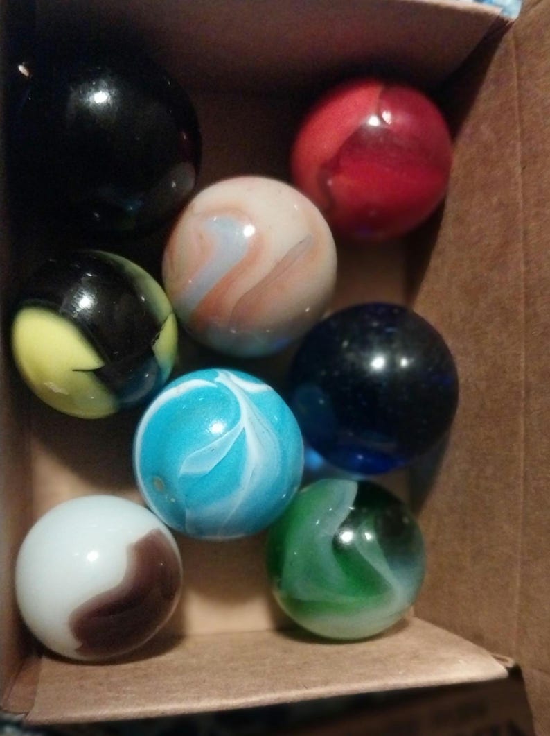 May include: A collection of colorful glass marbles in a cardboard box. The marbles are various colors, including black, red, blue, green, white, and a marbled blue and white. Some marbles have swirling patterns.