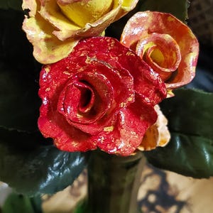 May include: Three artificial roses in a green glass vase. The roses are yellow, red, and orange with a glossy finish.