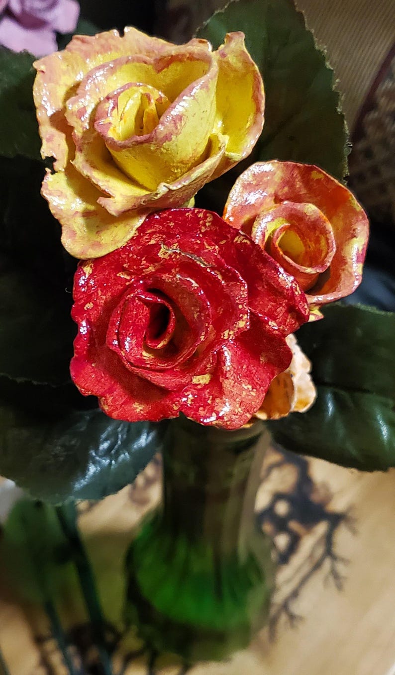 May include: Three artificial roses in a green glass vase. The roses are yellow, red, and orange with a glossy finish.