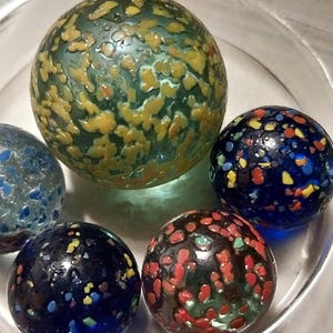 Op de afbeelding: Een verzameling van vijf kleurrijke glazen knikkers met gevlekte patronen. De knikkers bevinden zich in een glazen kom. De knikkers zijn blauw, groen, rood en geel.