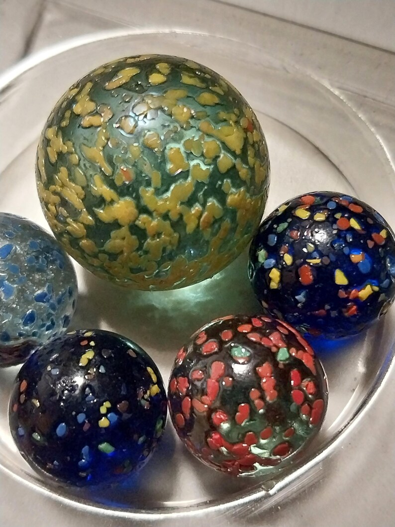 Op de afbeelding: Een verzameling van vijf kleurrijke glazen knikkers met gevlekte patronen. De knikkers bevinden zich in een glazen kom. De knikkers zijn blauw, groen, rood en geel.