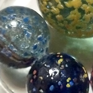 Op de afbeelding: Drie glazen knikkers met kleurrijke stippen. De knikkers zijn blauw, groen en zwart met gele, witte en rode stippen.