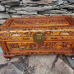 May include: A wooden chest with intricate carvings, featuring a detailed relief of ships and landscapes. The chest has a warm, reddish-brown hue, a brass clasp, and is set on small, decorative feet. The lid and sides are adorned with floral and geometric patterns.