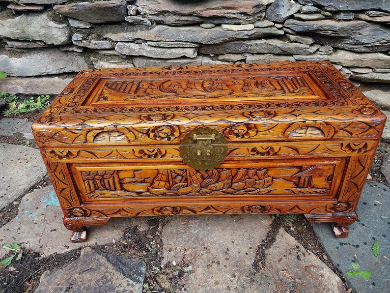 May include: A wooden chest with intricate carvings, featuring a detailed relief of ships and landscapes. The chest has a warm, reddish-brown hue, a brass clasp, and is set on small, decorative feet. The lid and sides are adorned with floral and geometric patterns.