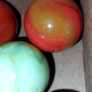 May include: Close-up of several colorful glass marbles. The marbles have swirling patterns in shades of red, orange, green, and brown. The marbles are in a cardboard box, suggesting they are for sale or storage.