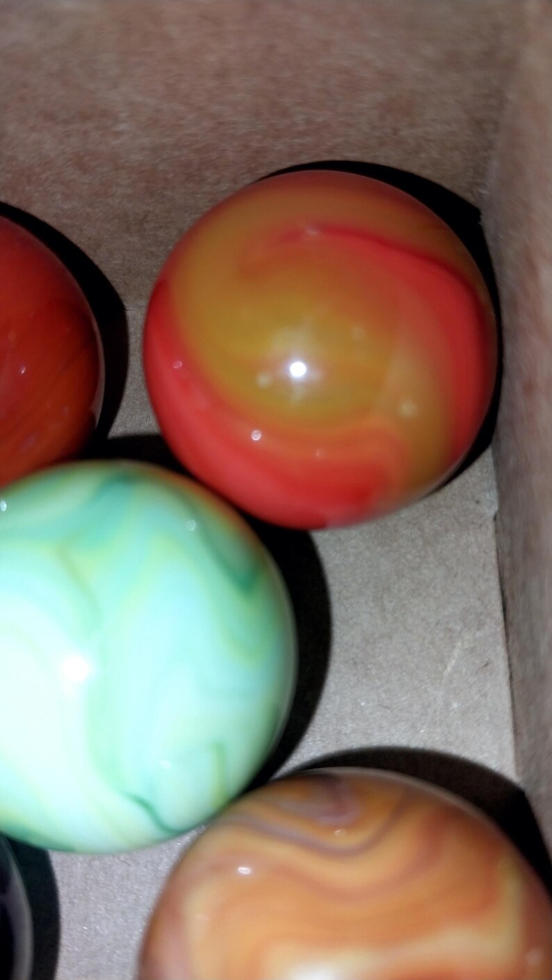 May include: Close-up of several colorful glass marbles. The marbles have swirling patterns in shades of red, orange, green, and brown. The marbles are in a cardboard box, suggesting they are for sale or storage.