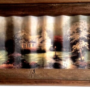 May include: A framed landscape print featuring a church with a steeple, a bridge, and trees. The print is set within a rustic, weathered wooden frame, creating a vintage aesthetic. The artwork depicts a serene outdoor scene.