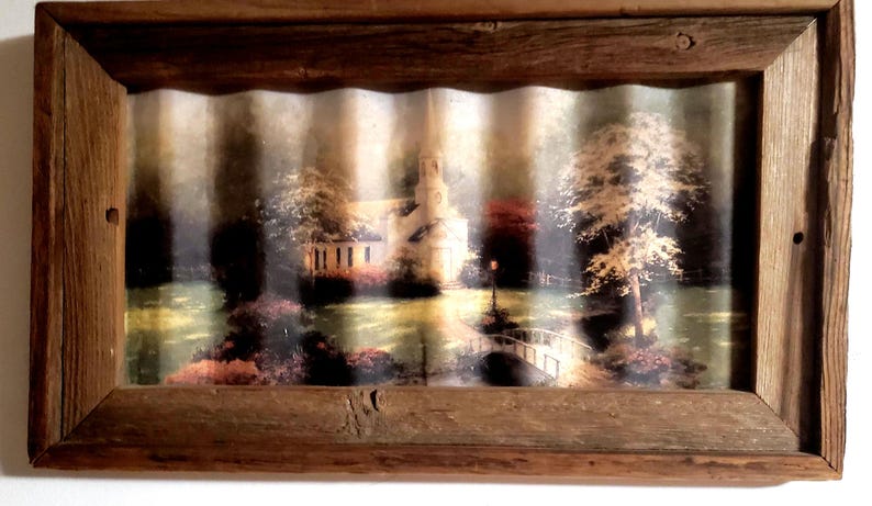 May include: A framed landscape print featuring a church with a steeple, a bridge, and trees. The print is set within a rustic, weathered wooden frame, creating a vintage aesthetic. The artwork depicts a serene outdoor scene.
