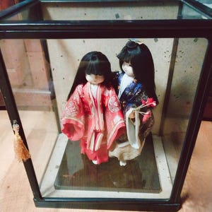 May include: Two Japanese dolls in a glass display case. The doll on the left wears a red kimono with a floral pattern, while the other doll wears a blue and white kimono. Both dolls have black hair and are standing on a wooden base.