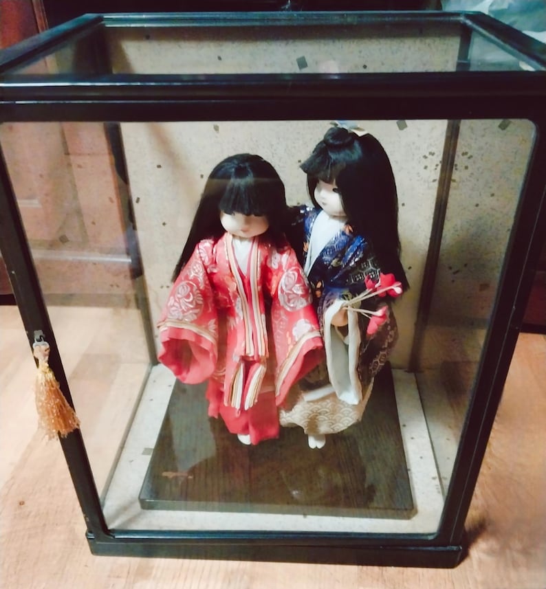 May include: Two Japanese dolls in a glass display case. The doll on the left wears a red kimono with a floral pattern, while the other doll wears a blue and white kimono. Both dolls have black hair and are standing on a wooden base.