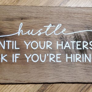 May include: A rectangular wooden sign with the words "hustle UNTIL YOUR HATERS ASK IF YOU'RE HIRING" in white script. The sign is made of distressed wood with visible grain and knots.