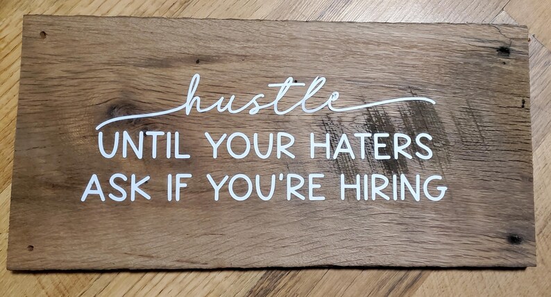 May include: A rectangular wooden sign with the words "hustle UNTIL YOUR HATERS ASK IF YOU'RE HIRING" in white script. The sign is made of distressed wood with visible grain and knots.
