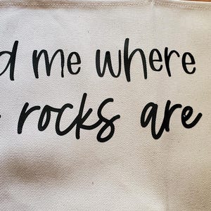 May include: A light beige canvas tote bag with the phrase "find me where the rocks are" printed in black cursive lettering. The bag has a simple design and a neutral color scheme.