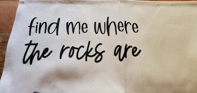 May include: A light beige canvas tote bag with the phrase "find me where the rocks are" printed in black cursive lettering. The bag has a simple design and a neutral color scheme.