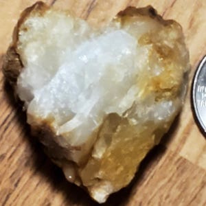 May include: A heart-shaped quartz crystal with a rough, textured surface. The crystal is primarily white with areas of tan and brown. A US quarter is next to the crystal for size comparison.