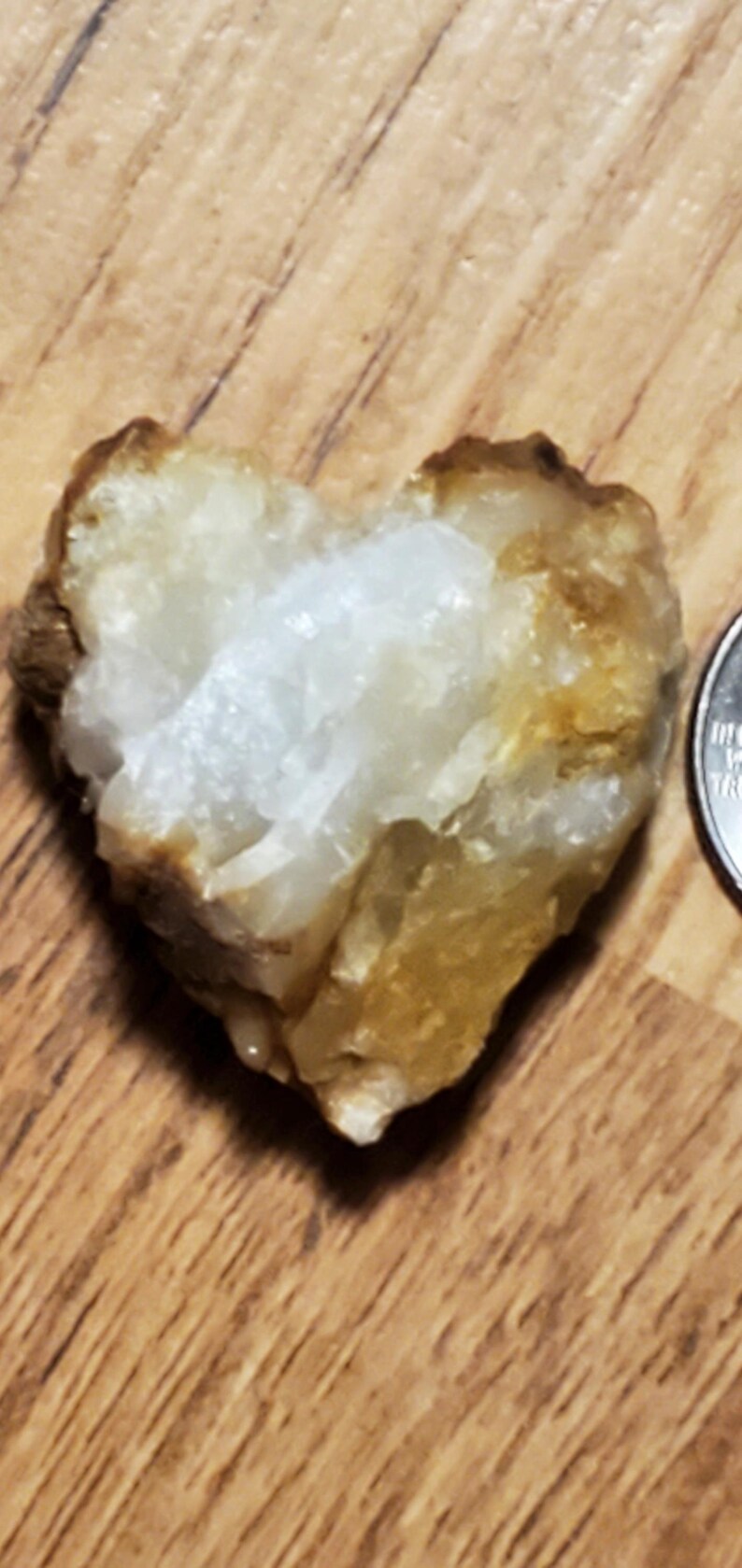 May include: A heart-shaped quartz crystal with a rough, textured surface. The crystal is primarily white with areas of tan and brown. A US quarter is next to the crystal for size comparison.
