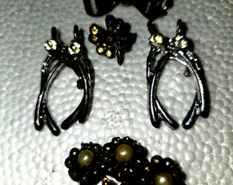 Op de afbeelding: Een verzameling van vijf metalen sieraden, waaronder een donkere strik, twee zilverkleurige wichelroede-vormige bedels met heldere kristallen, een kleine bloemvormige bedel met gele kristallen en een bloemenbroche met drie parels en bladeren.