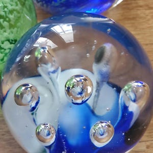 May include: A clear glass paperweight with blue and white swirls and embedded air bubbles. The spherical paperweight has a glossy finish and reflects light. Other glass spheres are visible in the background.