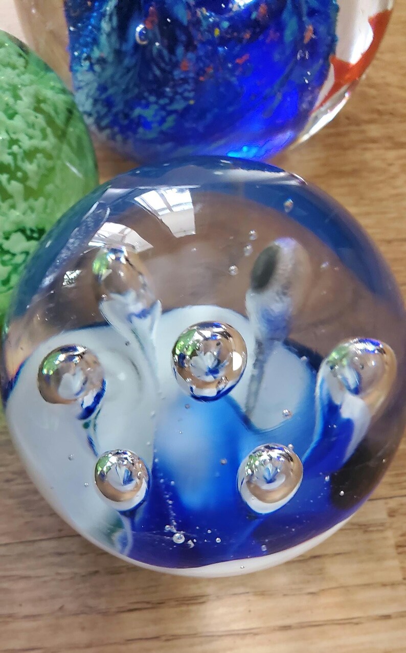 May include: A clear glass paperweight with blue and white swirls and embedded air bubbles. The spherical paperweight has a glossy finish and reflects light. Other glass spheres are visible in the background.