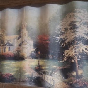 May include: A framed landscape print featuring a church with a steeple, a bridge, and trees. The artwork is set in a serene, natural environment with a muted color palette. The frame is made of wood.