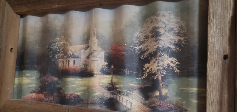 May include: A framed landscape print featuring a church with a steeple, a bridge, and trees. The artwork is set in a serene, natural environment with a muted color palette. The frame is made of wood.