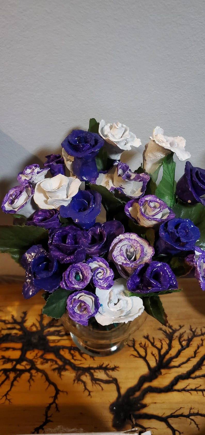 May include: A bouquet of purple and white paper roses in a clear glass vase. The roses are arranged in a cluster and have a textured, paper-like appearance.