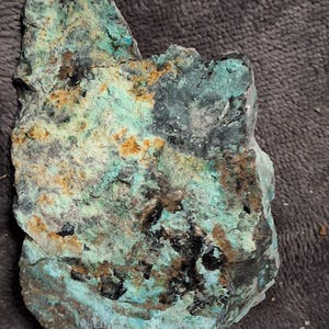 May include: A rough-hewn rock sample with a mix of colors. The stone features patches of turquoise, brown, black, and gray, creating a textured appearance. The rock's surface is uneven, with visible crevices and variations in color.