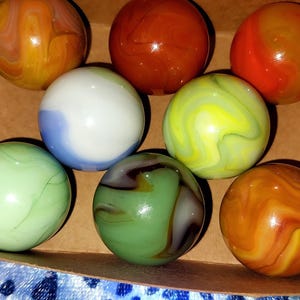 May include: A collection of colorful glass marbles in a cardboard box. The marbles feature swirls of orange, yellow, green, blue, and brown. The marbles are a classic toy, perfect for collectors or for playing games.